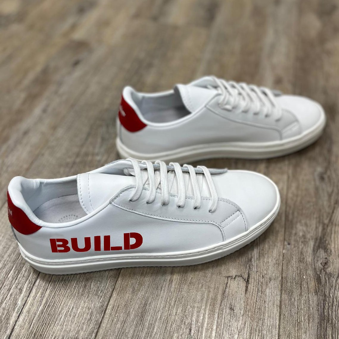 a pair of white sneakers with red heel and company logo on sides, placed on a wooden floor